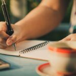 Manos de mujer escribiendo frases de autoestima en un cuaderno junto a una taza de café