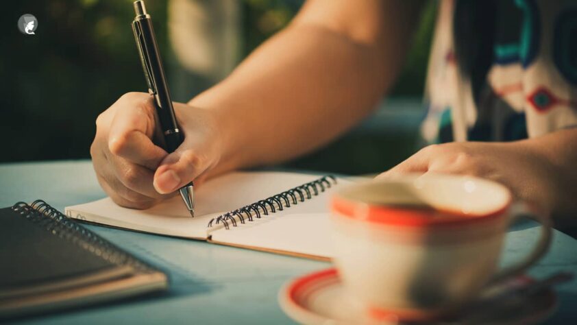Manos de mujer escribiendo frases de autoestima en un cuaderno junto a una taza de café